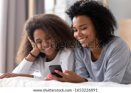 Happy african american mother and teen daughter using smartphone apps for shopping social media lying on bed, smiling black mom with teenage girl having fun watch videos on phone laughing in bedroom Royalty-Free Stock Photo #1653226891