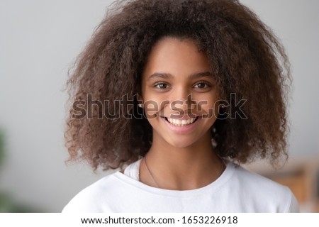 Head shot of smiling cute teenage african american girl with afro hair looking at camera at home, happy beautiful black teenager posing indoor, pretty adolescent mixed race teen school child portrait Royalty-Free Stock Photo #1653226918