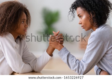Serious african american mom nanny and teen daughter arm wrestling looking in eyes fighting, angry black mother and teenage girl holding hands competing, family parent children disagreement concept