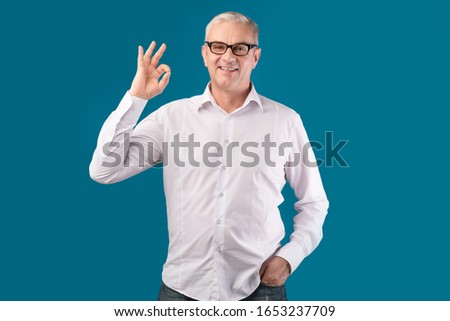 I'm Fine. Smiling senior man in specs showing ok gesture isolated over blue studio wall, free space