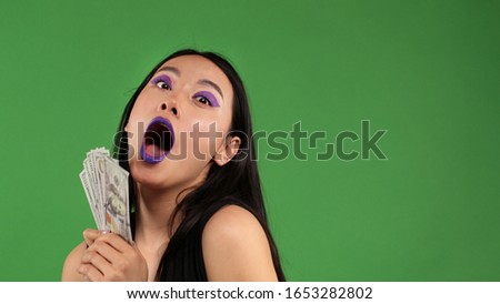 Asian hipster young woman with american dollars in hands on an isolated green background.Surprise, shock,Joyful emotions, fun, laughter and happiness.