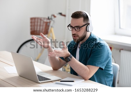 Focused young businessman in eyewear wearing headphones, holding video call with clients on laptop. Concentrated millennial man in glasses giving online educational class lecture, consulting customer. Royalty-Free Stock Photo #1653288229