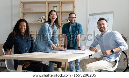 Portrait of excited young multiracial teammates, working together in modern office. Happy mixed race employees gathered near wooden table, enjoying workday time at workplace, looking at camera. Royalty-Free Stock Photo #1653288235