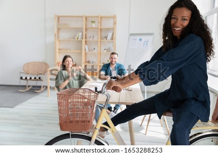 Focus on overjoyed young african american female employee, sitting on bicycle in office with blurred smiling colleagues on background. Happy biracial manager having fun, enjoying break pause time.