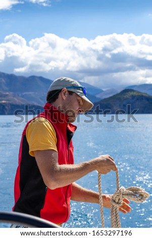 A man on a yacht is tying a knot on a rope, wearing a red yolk, wearing a cap and glasses. Vertical orientation.