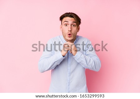 Young caucasian man posing in a pink background isolated praying for luck, amazed and opening mouth looking to front.