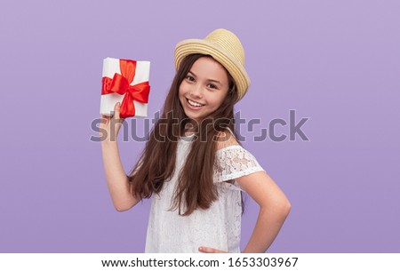 Cheerful little girl in white dress and hat holding white gift box with red ribbon and looking at camera while standing against lilac background