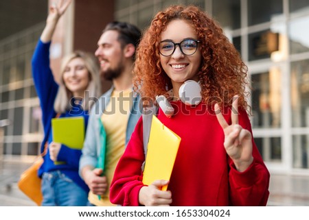 Happy young redhead female student in red blouse and eyeglasses holding paper fold and showing victory gesture, with fingers while standing with cheerful friends next to university building