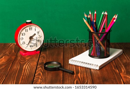 Colored pencils in a stand, notebook, alarm clock, magnifying glass on a wooden table.