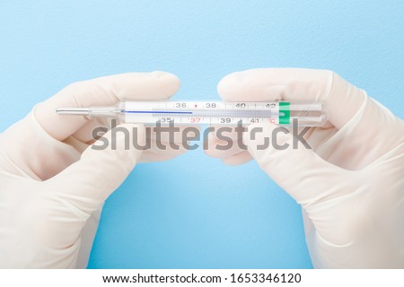 Doctor hands in white rubber protective gloves holding mercury thermometer on light blue table background. Pastel color. Closeup. Point of view shot. 