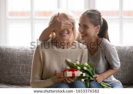 Joyful adult millennial daughter covering happy mommys eyes, holding flowers bunch, sitting together on sofa. Carrying young woman prepared surprise gift for mature mother for birthday celebration.