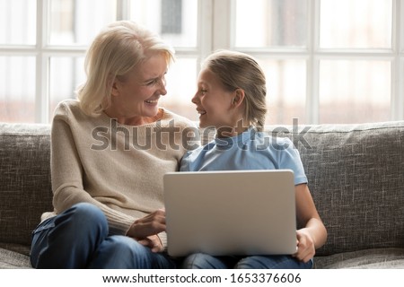 Overjoyed middle aged retired granny and happy school granddaughter sitting together on couch, holding laptop, watching funny movies or photos, feeling joyful, enjoying spending tome together.