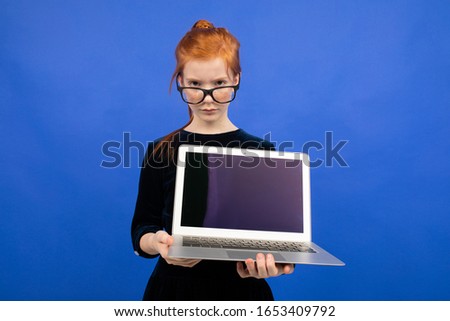 smart serious red-haired teenager girl in glasses holds a laptop with a blank screen mock up to insert a page on a blue background