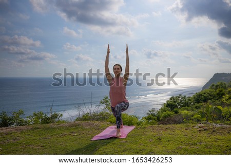 Young woman practicing yoga, standing in Virabhadrasana pose with raising arms. Outdoor yoga on the cliff. Warrior pose. Work out. Healthy lifestyle. Ocean in the background. Yoga retreat, Bali.