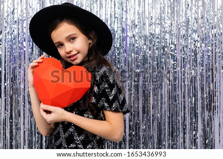 european cute girl in holiday dress and hat holds a red paper heart on a shiny background