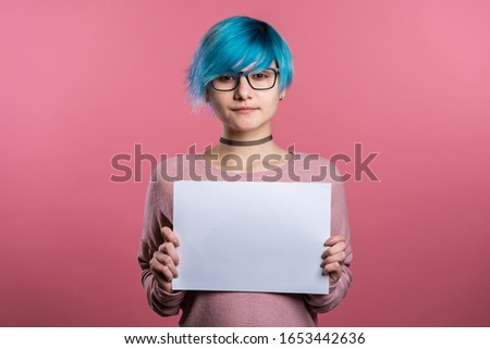Pretty girl with blue hair holding white a4 paper poster. Copy space. Smiling trendy woman on pink studio background.