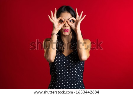 Positive girl making OK sign over eyes like glasses on red background and smiles to camera. Body language. Young woman.