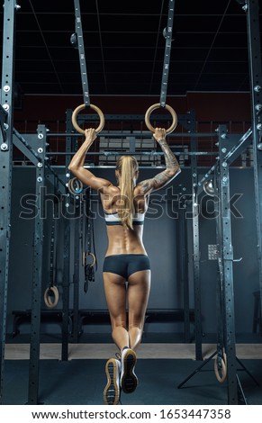 Muscular back of a girl Crossfit athlete in black sportswear is hanging on gymnastic rings with his back to the camera. Work out in the gym. Caucasian woman with long blond hair training olympic ring.