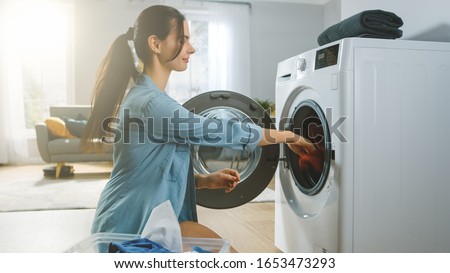 Beautiful Smiling Brunette Young Woman Sits in Front of a Washing Machine in Homely Jeans Clothes. She Loads the Washer with Dirty Laundry. Bright and Spacious Living Room with Modern Interior. Royalty-Free Stock Photo #1653473293