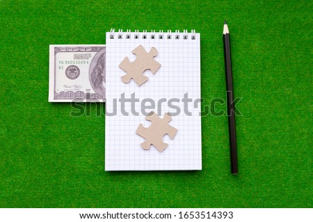 Notepad, two puzzle pieces, American dollar bill, pencil on a green background. 