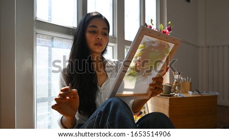 An artist girl sitting and lean on windows while looking on drawing canvas.