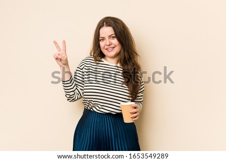 Young curvy woman holding a coffee joyful and carefree showing a peace symbol with fingers.