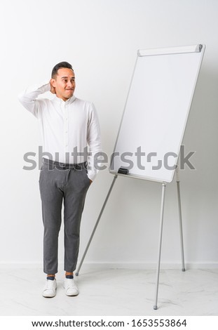 Young latin coaching man with a whiteboard isolated touching back of head, thinking and making a choice.
