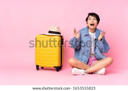 Young traveler Vietnamese woman with suitcase sitting on the floor over isolated pink background with surprise facial expression