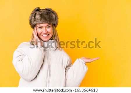 Young caucasian woman wearing a winter clothes isolated holds copy space on a palm, keep hand over cheek. Amazed and delighted.