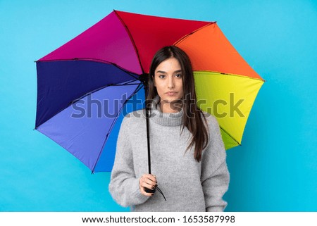 Young brunette woman holding an umbrella over isolated blue wall keeping arms crossed
