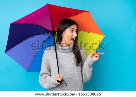 Young brunette woman holding an umbrella over isolated blue wall pointing finger to the side