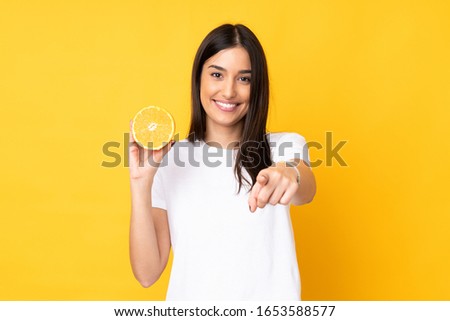 Young caucasian woman holding an orange isolated on yellow background points finger at you with a confident expression