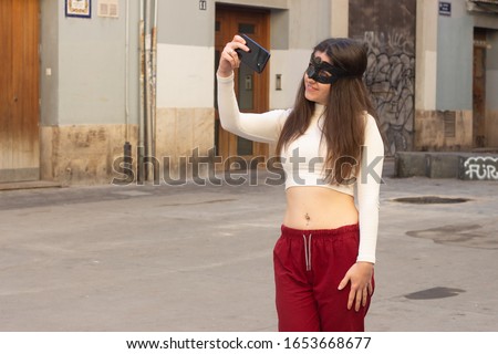 Portrait of attractive caucasian young woman with mask taking selfie, in the city, white top and red pants, dark air. Place for your text in copy space.