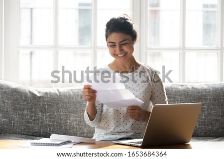 Indian ethnicity woman sitting on couch at home reading paper notice receive good news feels happy, cheerful student female looking at document enjoy exam results or college admission letter concept Royalty-Free Stock Photo #1653684364
