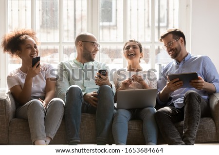 Sitting on sofa four multiracial friends using diverse devices hold phones laptop and tablet have fun together chatting enjoy live communication, generation and modern gadgets, social networks concept Royalty-Free Stock Photo #1653684664