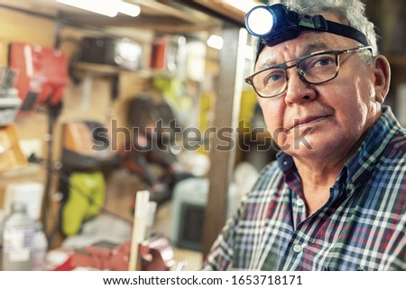 Senior craftsman portrait with headlamp led torch sitting in workhop with tools and equipment background. Old mature gray handyman tutor with eyeglasses. Experienced pensive person thinking new idea