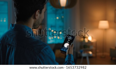 Young Handsome Man is Holding Smartphone with Active Smart Home Application. Person is Tapping the Screen to Turn On/Off the Lights in the Room. It's Cozy Evening in the Apartment.