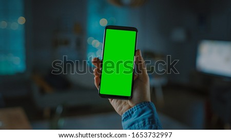 Close Up Shot of a Smartphone with Green Screen Great for Mock-up. Person is Holding Phone and Giving a Voice Command to Switch on the Light on the Backgroung in Living Room.