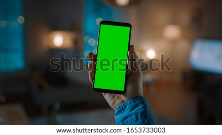 Close Up Shot of a Smartphone with Green Screen Great for Mock-up. Person is Holding Phone and Giving a Voice Command to Switch on the Light on the Backgroung in Living Room.