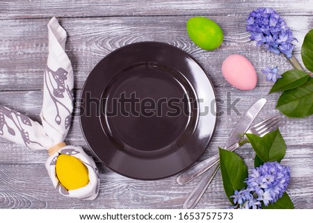 Easter composition with an empty black plate and an egg wrapped in a napkin in the form of rabbit ears, cutlery, colorful Easter eggs, spring flowers on a wooden gray table. Flat lay.
