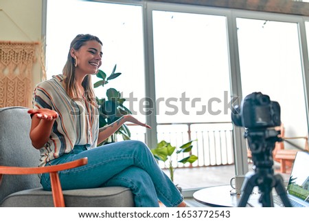 Attractive young woman blogger dressed in jeans and a blouse is recording on the camera while sitting on a comfortable chair at home.