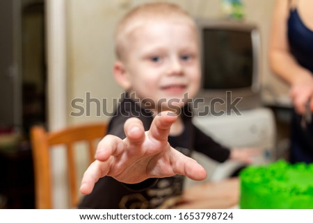 A little boy sitting at the table with a cake and pulls his hand to the camera.