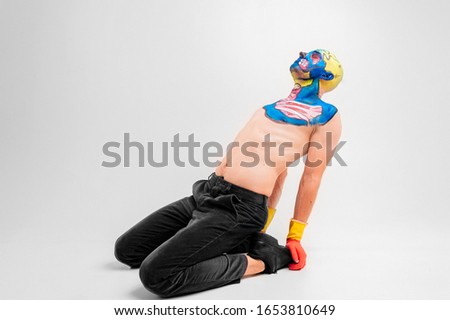 A strange man with makeup on his head and shoulder in red-yellow gloves, black pants and boots, no T-shirt sits on a floor