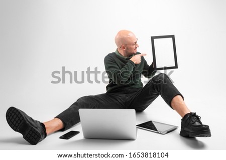 Handsome caucasian male holds a phone in one hand, tablet in other hand with laptop, sits on a floor isolated on white background