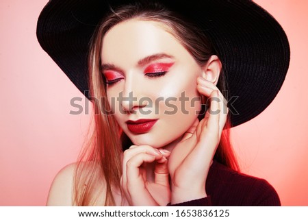 Gorgeous blonde in a big hat with burgundy make-up, red lips, and a burgundy dress. Girl in burgundy earrings, portrait of a model. Fashion photo of bright red makeup, model on a red background