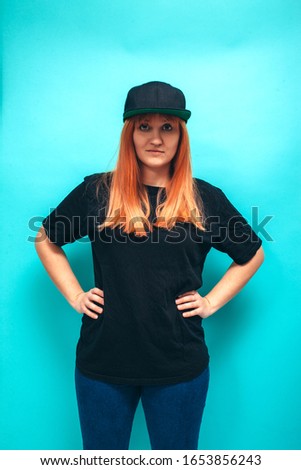 Handsome hipster female with bright red hair wears black oversized t shirt and jeans, isolated on blue studio wall.