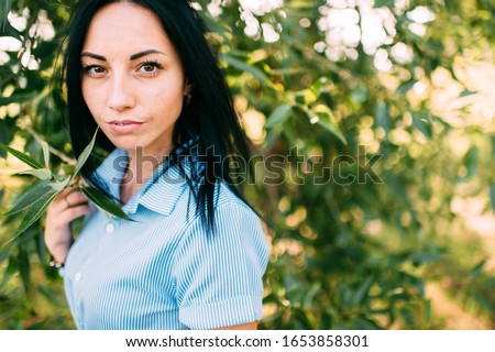 Lovely young caucasian girl with big brown eyes, long dark hair and perfect smile in short blue dress near the green tree in the garden, portrait isolated on green background