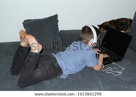 kid, a boy of primary school age in headphones, jeans, a blue shirt lies on a sofa in front of an open laptop with black, a cat sits nearby and looks, concept children and the Internet