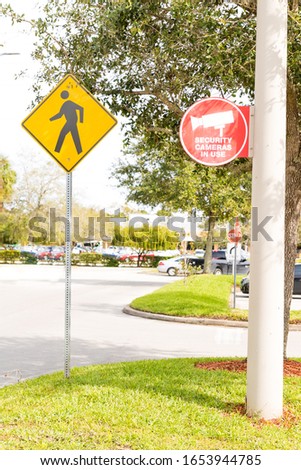 A yellow crosswalk sign and a red "SECURITY CAMERA IN USE" sign in a shopping plaza parking lot.