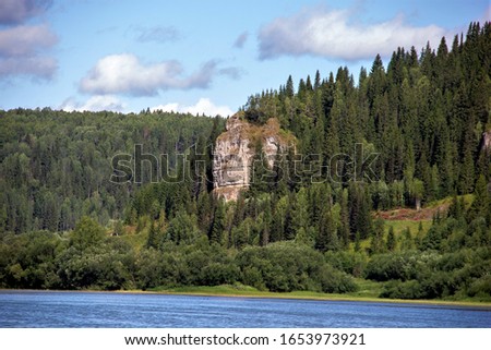 grey rocky slope of the mountain surrounded by green forest on a summer day under blue sky with clouds